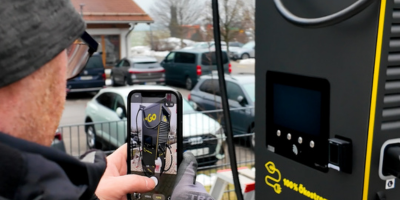 Person mit Mütze und Handschuhen fotografiert mit dem Smartphone eine E-Ladestation; im Hintergrund stehen mehrere Autos auf einem Parkplatz.